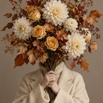Beeld van een persoon in een crèmekleurige wollen jas die een groot herfstboeket met dahlia's, rozen en bessen voor het gezicht houdt.