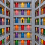 Afbeelding van een symmetrische betonnen gevel met balkons en felgekleurde deuren, met één klein figuur op het middelste balkon.