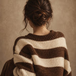 Portret van een vrouw van achteren met een knotje en een grove bruin-crème gestreepte trui voor een bruine achtergrond.