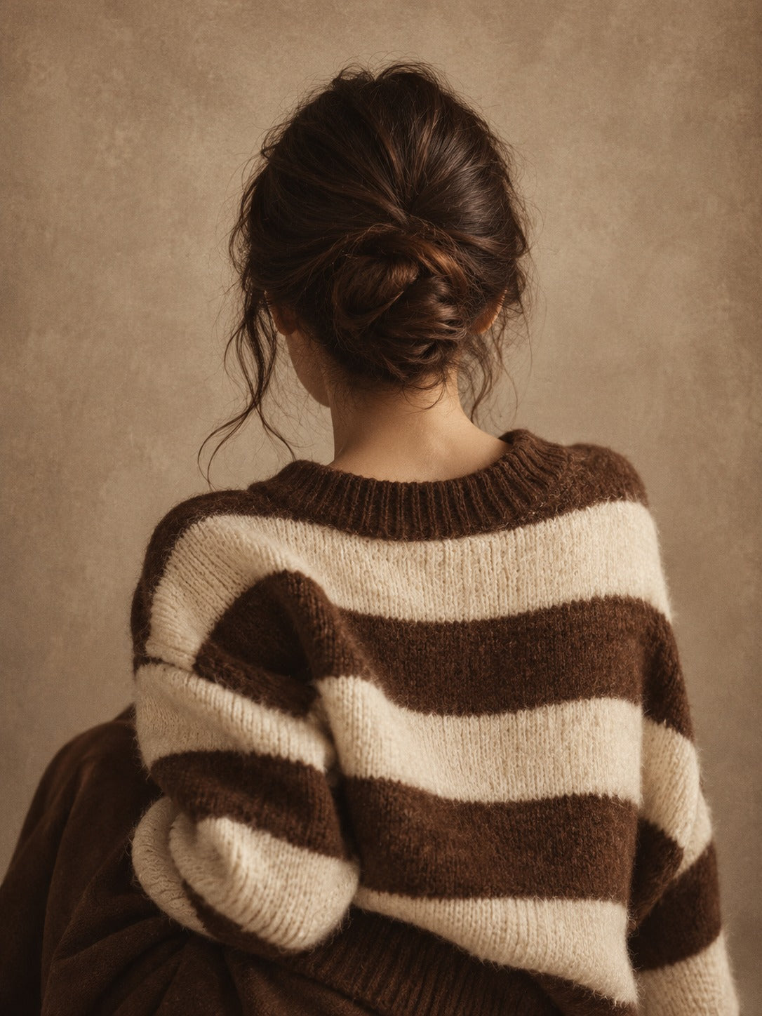 Portret van een vrouw van achteren met een knotje en een grove bruin-crème gestreepte trui voor een bruine achtergrond.