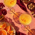Bovenaanzicht van twee coupeglazen met gouden drankje, schaaltjes met noten, sinaasappelpartjes en bordeaux bloemen op een roze tafel.