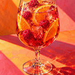 Wijnglas met rood drankje, frambozen, ijsblokjes en sinaasappelschijven tegen een achtergrond van oranje en roze lichtstrepen.